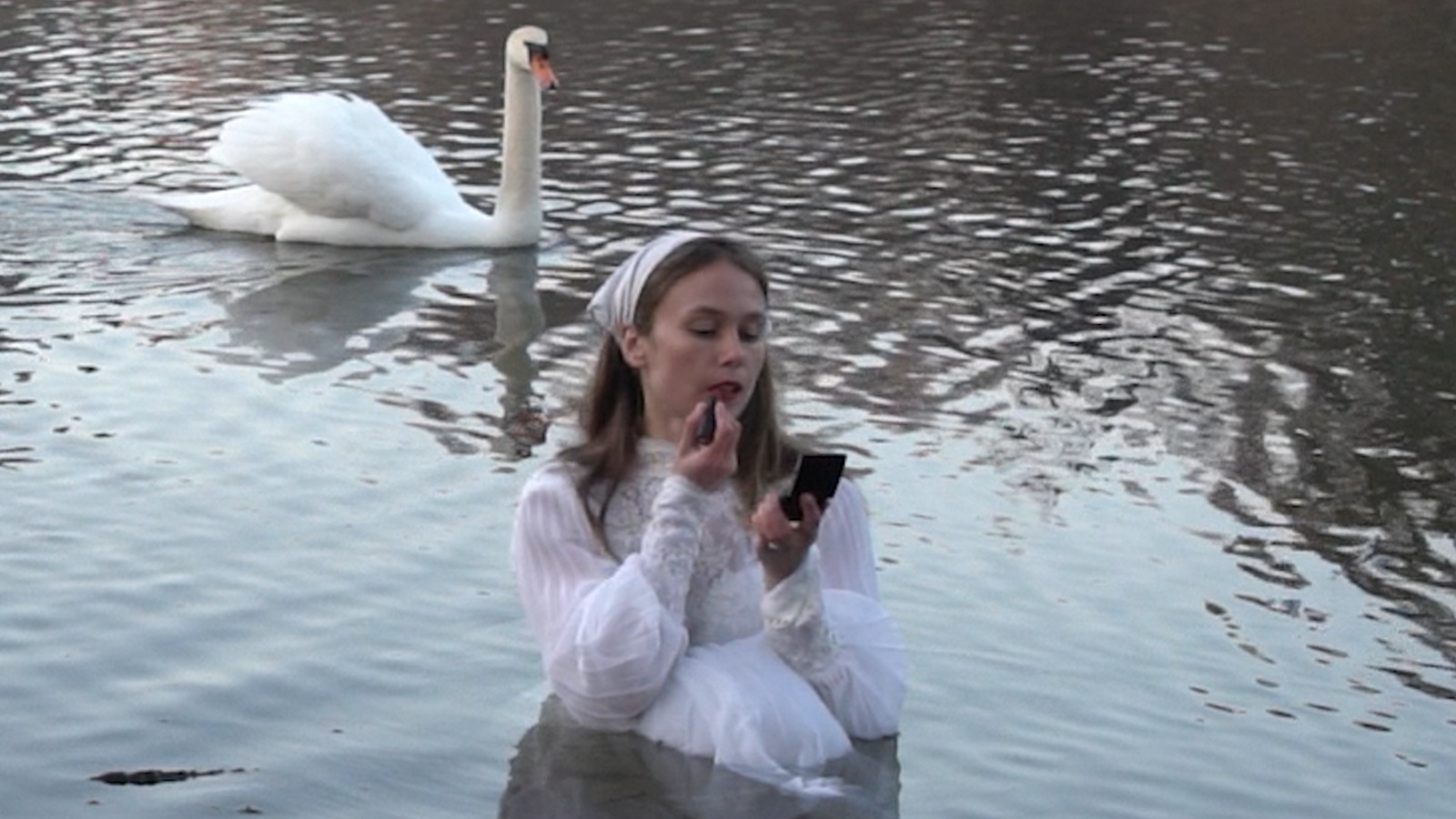 Photo by Sage Szkabarnicki-Stuart, of her standing in water and applying lipstick, with a swan in the background